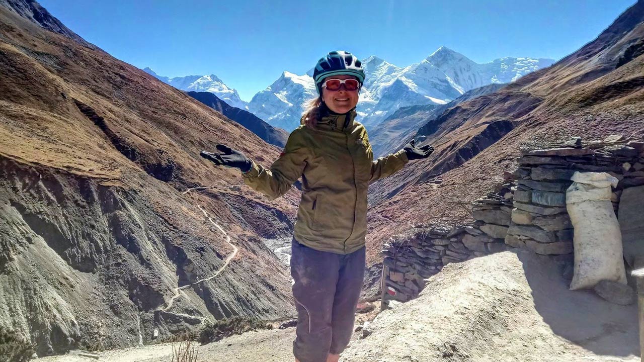 A biker in her green jacket expresses her joy stretching her hands sideways while taking a picture with the view of Annapurna and Gangapurna mountains.