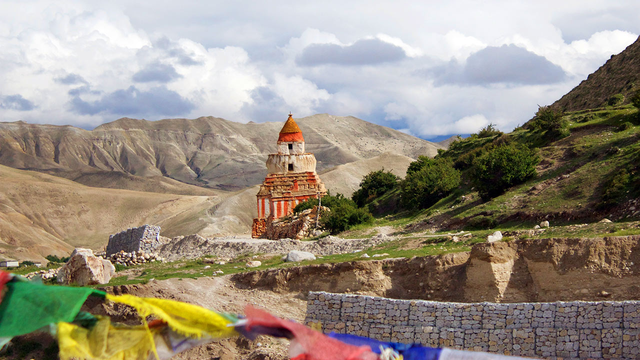 A stupa painted in red and white stands tall in the mystic land of Upper Mustang with the barren hill on the background and the greeneries on the side.